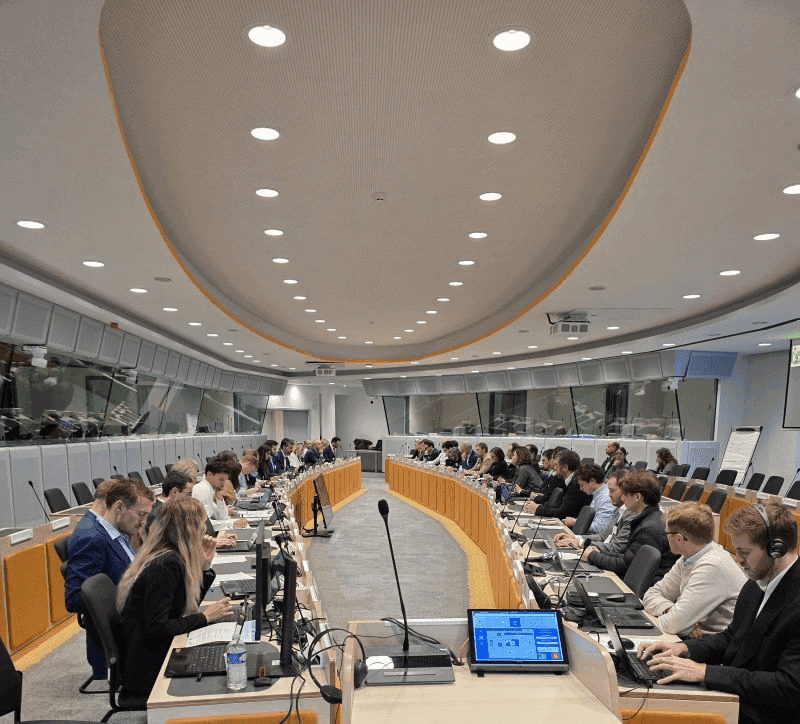 Conference room filled with people seated at a large, curved table, using laptops. Bright ceiling lights illuminate the space, creating a focused atmosphere.